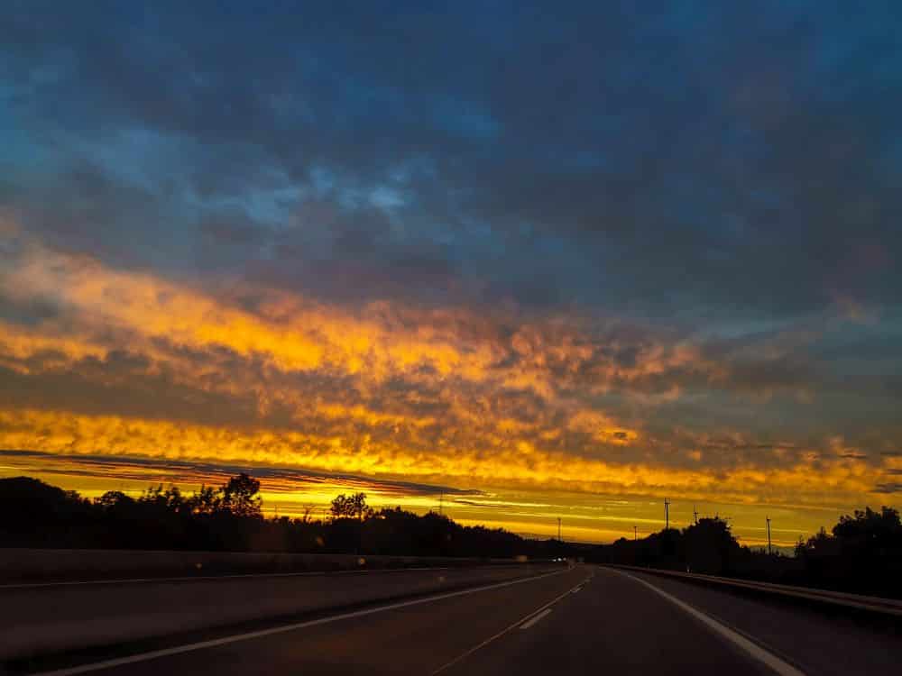 Ein lebhafter schwäbischer Sonnenuntergang mit orangefarbenen und gelben Wolken erhellt den Himmel über einer leeren Autobahn, die von Silhouetten von Bäumen und Strommasten begrenzt wird.