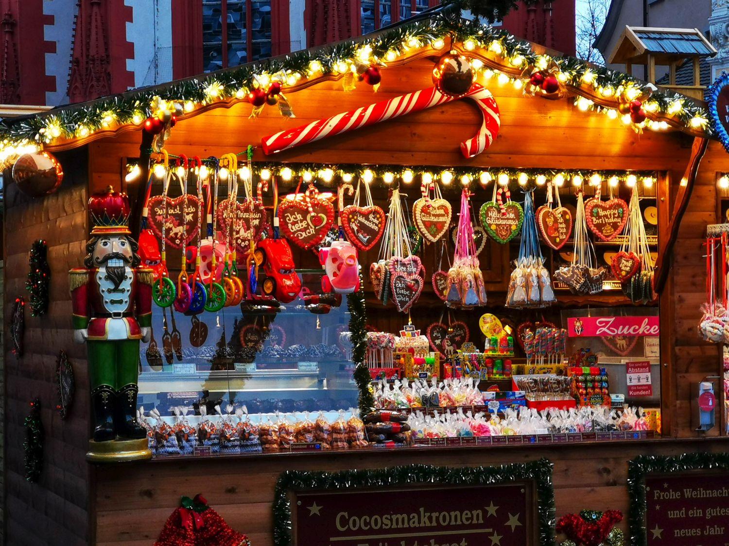 Ein festlicher Weihnachtsmarktstand aus Holz, geschmückt mit Lichtern, Zuckerstangen und einem Nussknacker, bietet bunte Süßigkeiten, herzförmige Kekse, Schokolade und weihnachtliche Leckereien.