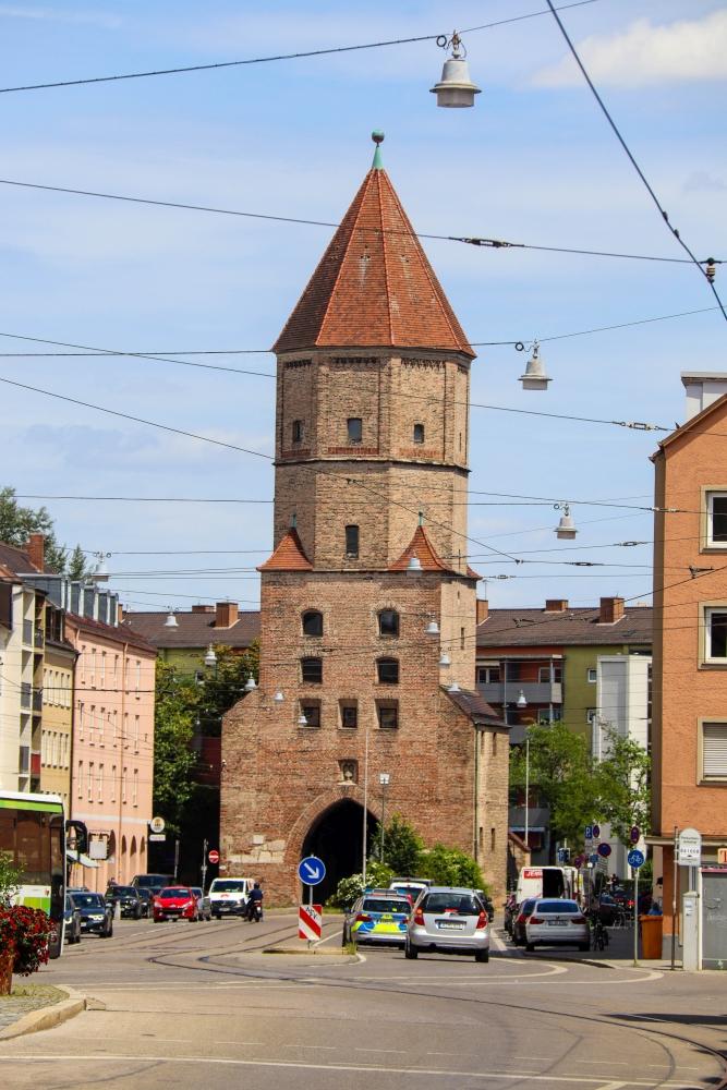 Ein historischer Stadttorturm aus Backstein mit rotem Spitzdach steht an einer belebten Augsburger Kreuzung, umgeben von modernen Gebäuden, Autos und Straßenlaternen unter einem teilweise bewölkten Himmel.