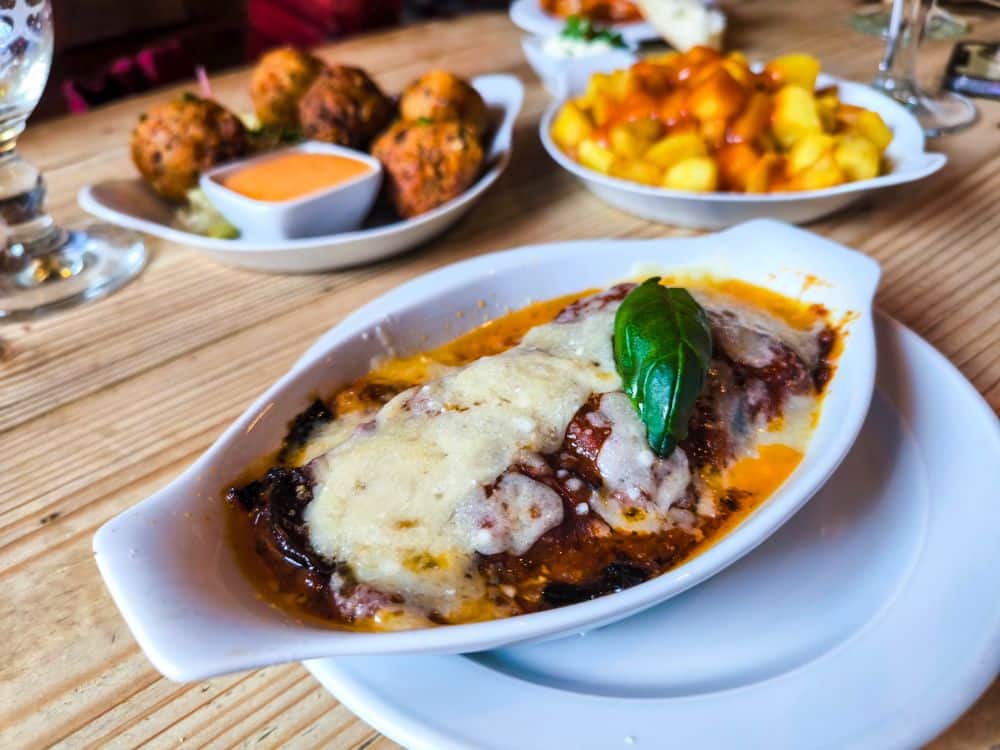 Ein weißes Gericht aus gebackenem Auberginen-Parmesan, garniert mit geschmolzenem Käse und einem Basilikumblatt, serviert zusammen mit gelben Kartoffeln, Soße und frittierten Vorspeisen auf einem Holztisch in Mechelen.