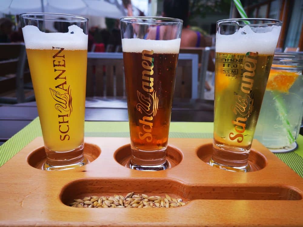Drei Schwanen-Biergläser, jedes mit einer anderen Sorte Ehinger Bier gefüllt, stehen auf einem Holztablett, in dessen Fach Gerste eingelegt ist. Jedes Glas zeigt das ikonische Schwanen-Logo. Im Hintergrund trägt ein erfrischendes Getränk mit Strohhalm zur lebhaften Szene bei.