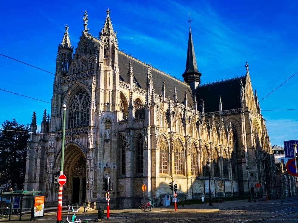 Kirche im gotischen Stil mit kunstvollen Steinmetzarbeiten, Spitzbögen, großen Glasfenstern und einem hohen Kirchturm vor einem strahlend blauen Himmel an einem sonnigen Tag. Vor der Kirche sind Fahrräder und Straßenschilder zu sehen.