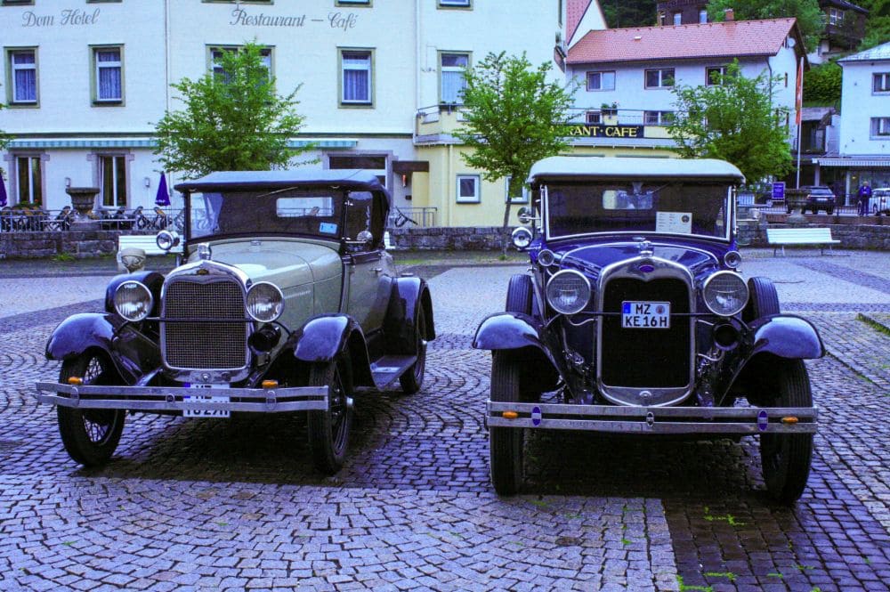 Zwei nebeneinander auf einer Kopfsteinpflasterstraße vor einem Gebäude mit der Aufschrift „Restaurant-Café“ geparkte Oldtimer vermitteln den Charme von St. Blasien. Das klassische Design der Autos wird durch Bäume und weitere Gebäude im Hintergrund ergänzt und schafft so eine zeitlose Szene.
