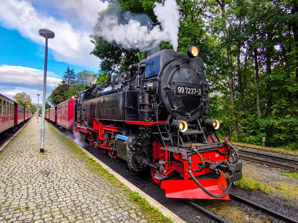 Eine alte schwarz-rote Dampflokomotive stößt weißen Rauch aus und steht auf einem Kopfsteinpflaster-Bahnsteig am Harzer Bahnhof. Der Zug, der aus roten Personenwagen besteht, ist von üppigen grünen Bäumen unter einem teilweise bewölkten Himmel umgeben. Deutschland