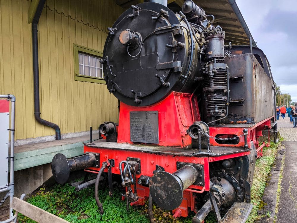 Eine alte Dampflokomotive, die an die Sauschwänzlebahn erinnert, steht stolz neben einem Holzgebäude. Ihr roter Sockel und die schwarzen Komponenten erwecken ein Gefühl von Nostalgie, mit verwitterten Details und Gras, das ihren Rahmen sanft vor einem bewölkten Himmel umgibt.