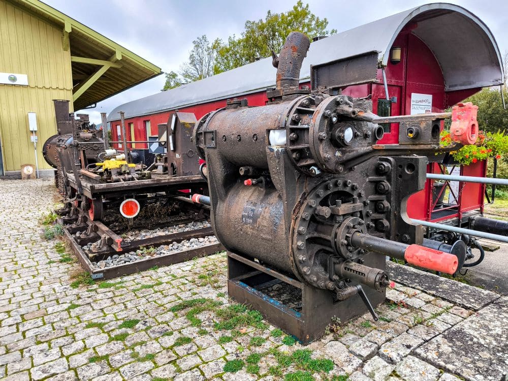 Eine große, verrostete Lokomotive steht auf einem Kopfsteinpflasterweg neben einem roten Oldtimer-Zugwagen und weckt ein Gefühl von Nostalgie. Im Hintergrund rahmen ein gelbes Gebäude und Bäume unter einem bewölkten Himmel dieses Schienen-Tableau in einem Freilichtmuseum oder einer Eisenbahnausstellung ein.