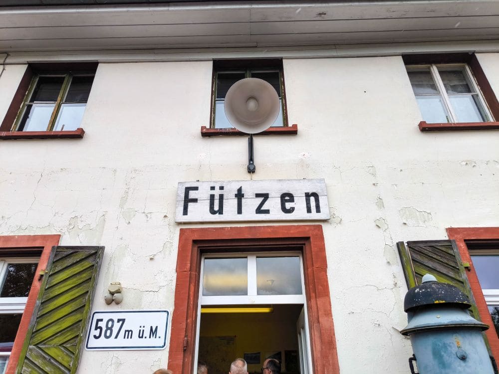 Das Bild zeigt das nostalgische Äußere eines Gebäudes mit einer abgenutzten weißen Fassade, an der sich zwei obere Fenster und ein großer Lautsprecher befinden. Über der Tür stehen Schilder mit der Aufschrift „Fützen“ und „587 m ü. M.“ – eine Haltestelle entlang der historischen Sauschwänzlebahn.