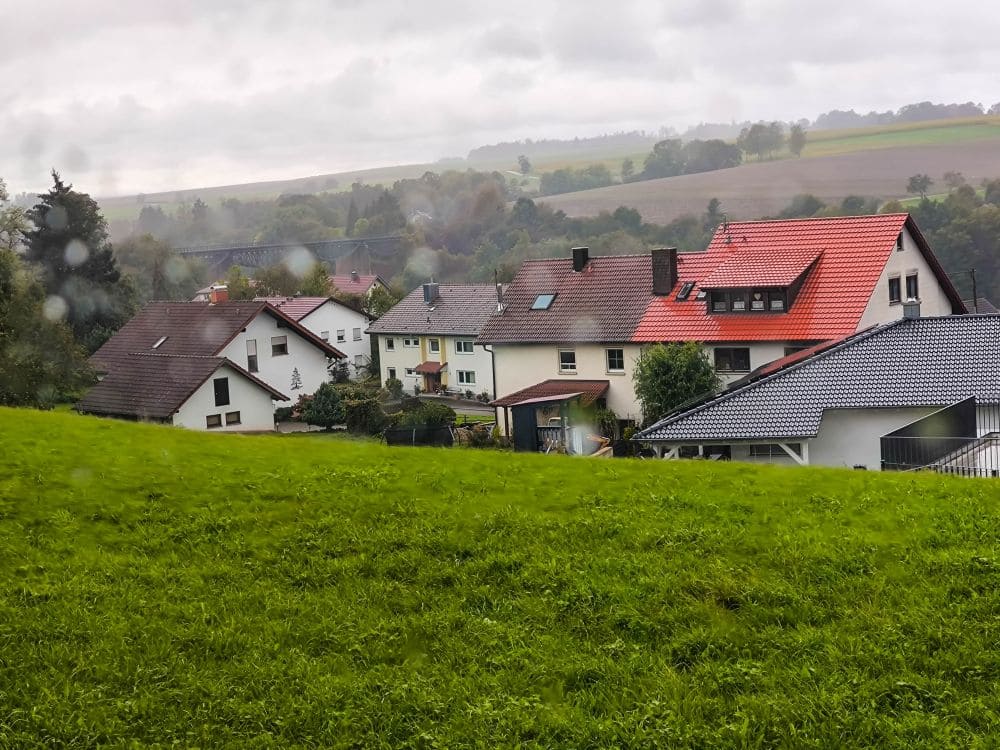 Eine ländliche Landschaft mit mehreren Häusern mit roten und grauen Dächern schmiegt sich an einen grasbewachsenen Hügel. In der Ferne zeichnen Schienen den Hintergrund unter einem wolkigen Himmel über weit entfernten Feldern nach und schaffen eine nostalgisch angehauchte, malerische Szene.