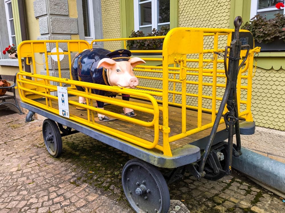 Eine Schweinestatue mit Schaffnermütze und -jacke steht auf einem gelben, alten Gepäckwagen, der an die Sauschwänzlebahn-Zeit erinnert. Der Wagen steht auf Kopfsteinpflaster vor einem Gebäude mit strukturierten Wänden und Topfblumen auf Fensterbänken.