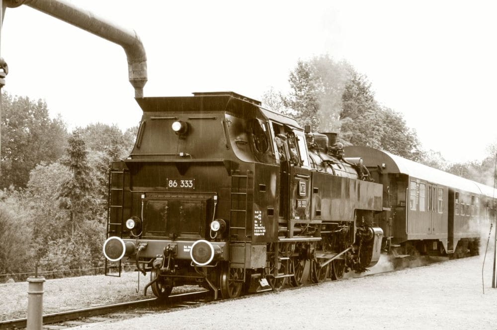 Eine historische Dampflok mit der Aufschrift „86 333“ auf der Vorderseite zieht einen Zug über die Schienen der Sauschwänzlebahn, eingehüllt in Dampf. Im Hintergrund sind Bäume zu sehen, und links ist ein Wasserfüllgerät zu sehen. Der Sepiaton trägt zur Nostalgie bei und verleiht dem Bild ein altmodisches Gefühl.