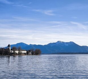 In einem ruhigen Chiemsee mit sanften Kräuselungen spiegeln sich eine Kirche und Bäume an seinem Ufer, während sich im Hintergrund schneebedeckte Berge unter einem klaren blauen Himmel mit einem schwachen Kondensstreifen erheben.