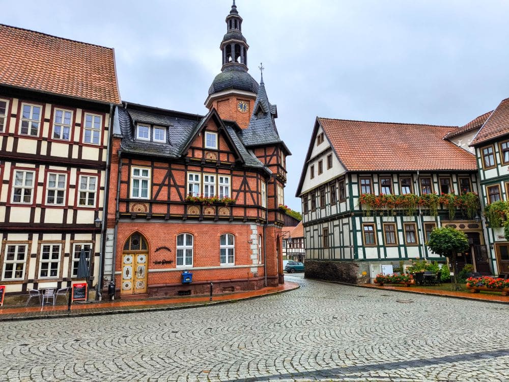 Das charmante Straßenbild in Stolberg zeichnet sich durch traditionelle Fachwerkhäuser mit steilen, roten Ziegeldächern aus. Eine Kopfsteinpflasterstraße schlängelt sich durch die Stadt, gesäumt von bunten Blumen und Grünflächen. Der Himmel ist bedeckt, was dieser malerischen Kulisse einen Hauch von Geheimnis verleiht.
