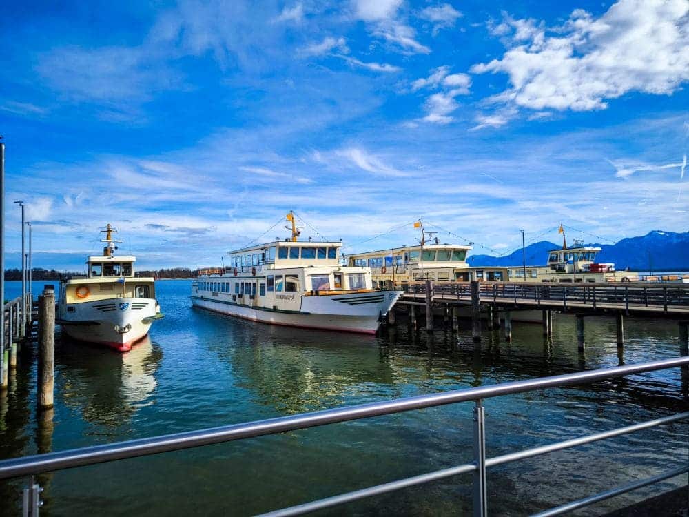 An einem klaren Tag liegen drei Fähren an einem Pier am Chiemsee, umgeben von ruhigem Wasser. Der Himmel ist strahlend blau mit vereinzelten Wolken. Im Hintergrund sind schneebedeckte Berge zu sehen, im Vordergrund ein Metallgeländer.