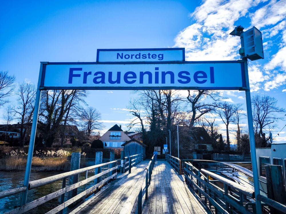 Ein Holzsteg erstreckt sich in das ruhige Wasser des Chiemsees und führt zu einer Insel mit einem einladenden Schild darüber, auf dem „Nordsteg Fraueninsel“ steht. Bäume und malerische Häuser säumen den Hintergrund unter einem strahlend blauen Himmel mit vereinzelten Wolken.