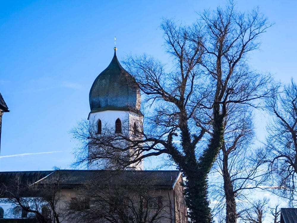 Vor dem klaren blauen Himmel ragt anmutig eine traditionelle bayerische Kirche mit einem zwiebelförmigen Turm empor. Um sie herum stehen blattlose Bäume mit kunstvoll gewundenen Ästen, die die Winterstimmung in der Nähe des Chiemsees noch verstärken.