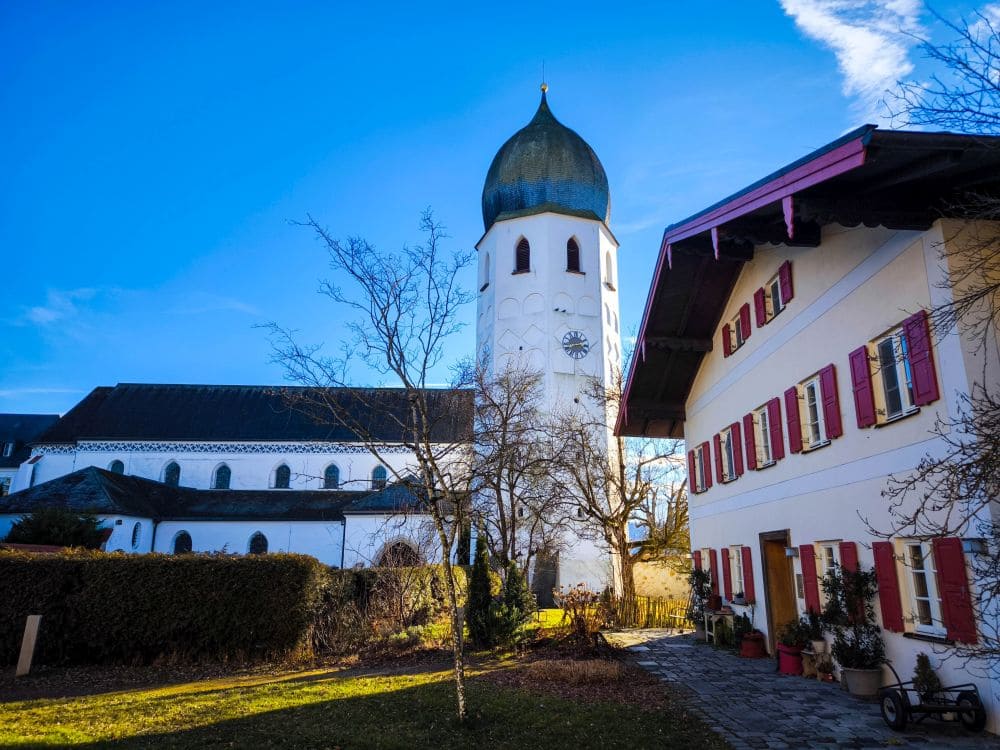 Eine malerische Aussicht auf ein charmantes Dorf in der Nähe des Chiemsees mit einem hohen Kirchturm mit Kuppel und Uhr. Der Himmel ist klar und blau. Im Vordergrund steht ein weißes Gebäude mit roten Fensterläden, umgeben von Bäumen und Grün.