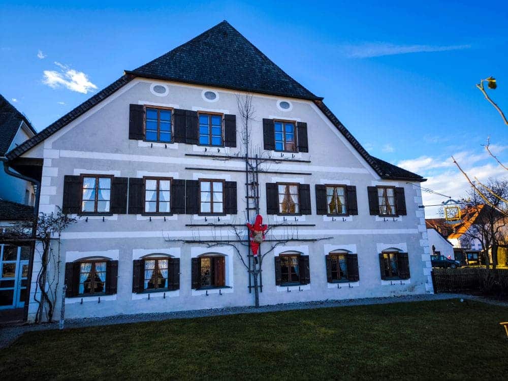 Ein traditionelles europäisches Haus mit weißen Wänden und dunklen Holzfensterläden bietet Aussicht auf die malerische Schönheit des Chiemsees. Die rote Silhouette einer kletternden Figur ziert die Fassade unter einem klaren Himmel, während sich im Vordergrund eine Grasfläche erstreckt.