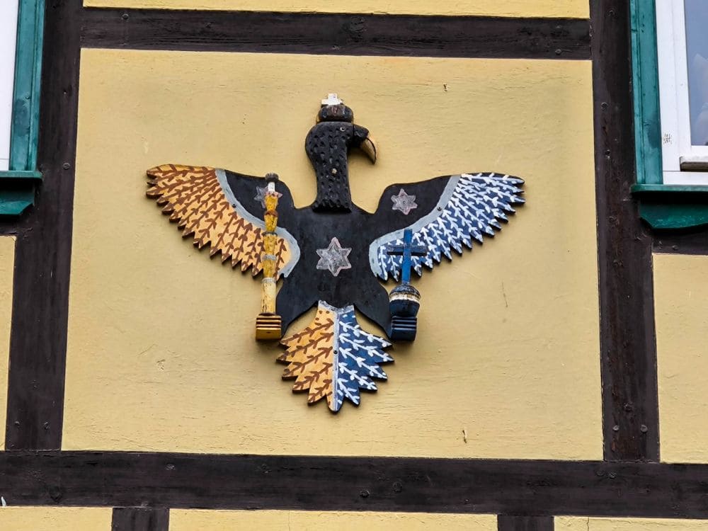 Ein dekoratives Adleremblem schmückt stolz eine gelbe Wand in Stolberg. Es zeigt einen schwarzen Adler mit blauen und weißen Flügeln auf der rechten Seite und goldenen und weißen auf der linken Seite, die jeweils einen Gegenstand umklammern. Sterne und aufwendige Kronendetails vervollständigen diese majestätische Darstellung.