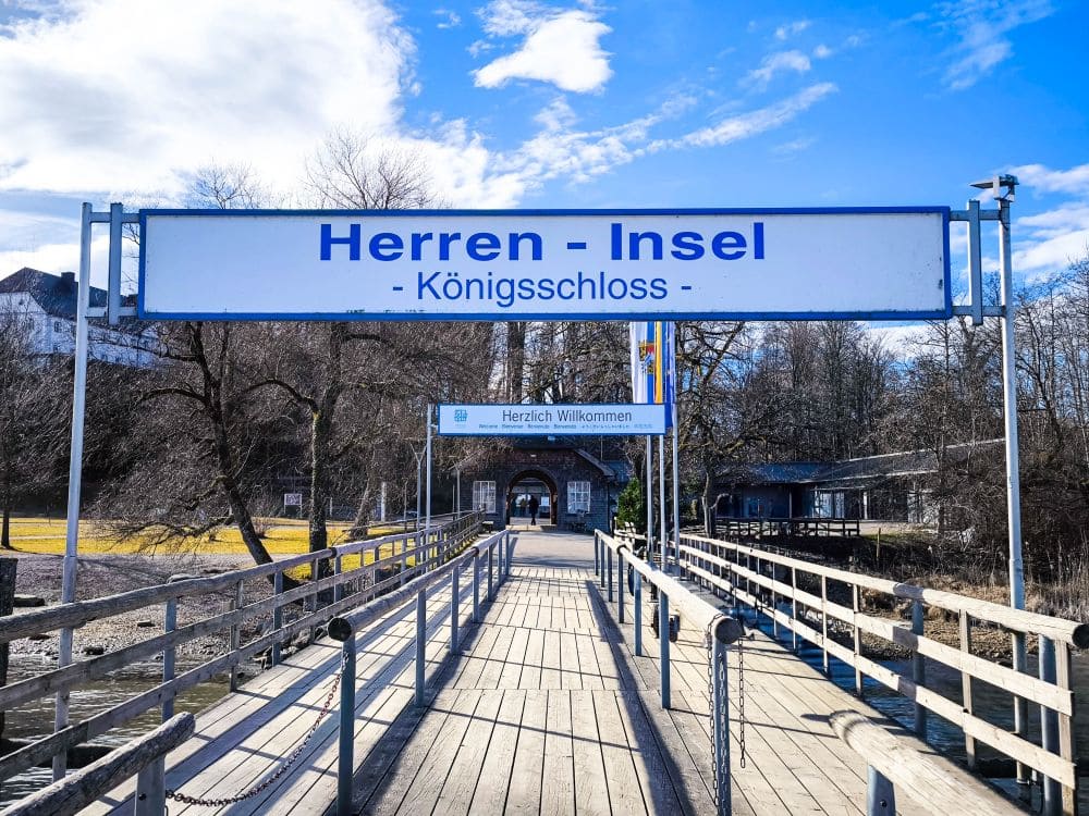 Blick auf den Eingang zur Herreninsel am Chiemsee mit Schildern mit der Aufschrift „Herren - Insel“ und „Königsschloss“ über einem Holzsteg. Die Szene zeigt Bäume, einen klaren blauen Himmel und Gebäude im Hintergrund und spiegelt die ruhige Schönheit dieses idyllischen Ortes perfekt wider.