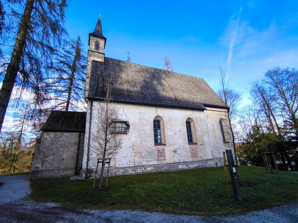 Eine kleine, historische Steinkirche mit Schrägdach und Glockenturm steht friedlich in der Nähe des Chiemsees, umgeben von kahlen Bäumen unter einem klaren blauen Himmel. Ein Kiesweg schlängelt sich anmutig um das Gebäude und unterstreicht seinen zeitlosen Charme.
