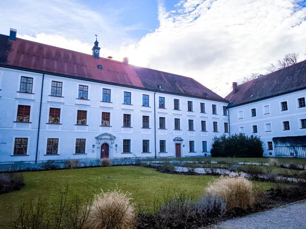 Das historische weiße Gebäude mit dem roten Ziegeldach, den zahlreichen Fenstern und den gewölbten Türen erinnert an den Charme des Chiemsees. Es ist von einem gepflegten Garten mit Gras und Sträuchern umgeben. Im Hintergrund ist ein teilweise bewölkter Himmel zu sehen.