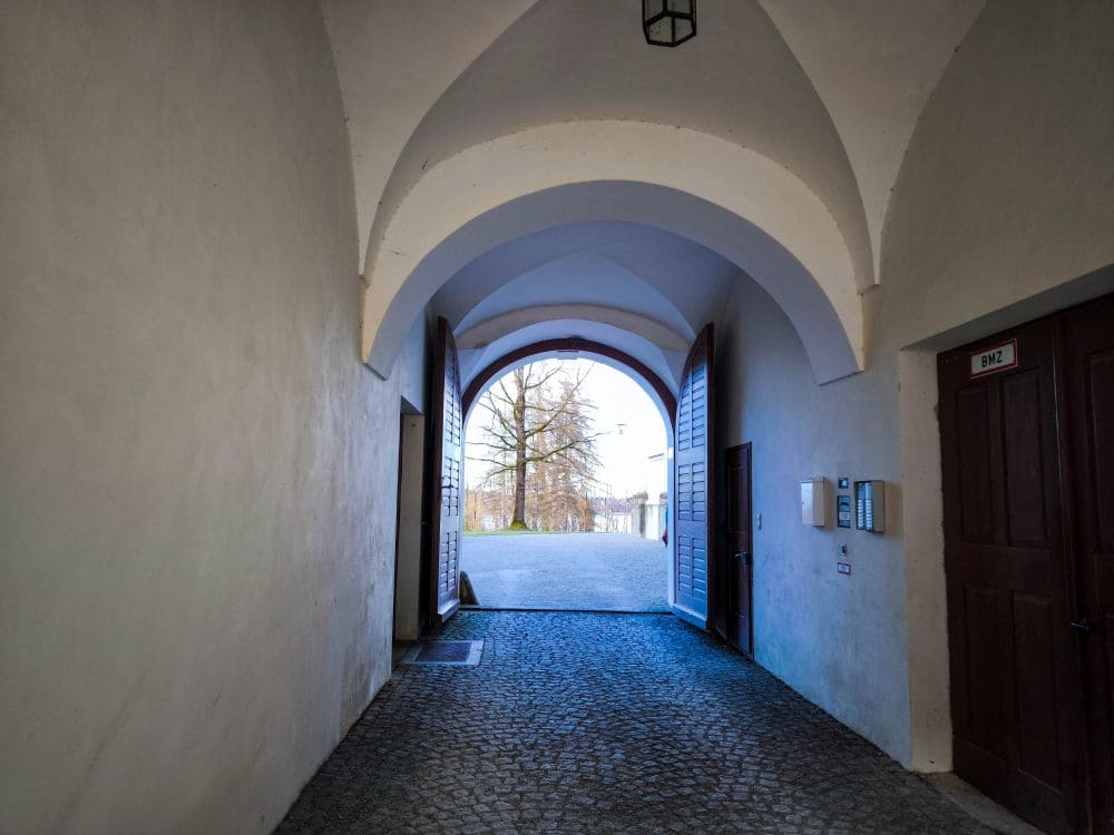 Das Bild zeigt einen Steintunnel mit gewölbter Decke, der zu einem offenen Holztor führt. Hinter dem Tor entfaltet sich ein gepflasterter Weg in Richtung der bezaubernden, von Bäumen gesäumten und in Sonnenlicht getauchten Chiemseeregion.