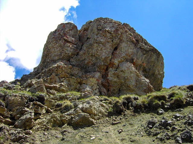 Eine große, schroffe Felsformation erhebt sich vor einem strahlend blauen Himmel mit vereinzelten Wolken. Im Vordergrund, in der Nähe der Plattkofelhütte, sind Grasflächen und unebenes Gelände zu sehen, was die natürliche, bergige Landschaft betont.