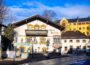 Eine malerische Szene eines charmanten Hotels in Bad Aibling zeigt traditionelle bayerische Architektur. Das Gebäude verfügt über kunstvolle Verzierungen, Holzbalkone und Wandmalereien. Bäume ohne Blätter und benachbarte Gebäude stehen stolz unter einem strahlend blauen Himmel.