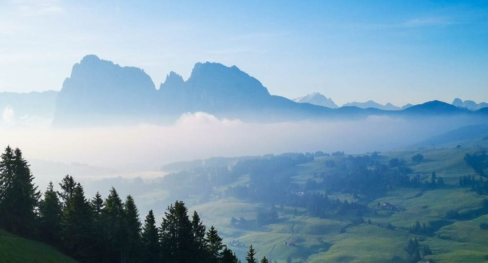 Eine ruhige Morgenlandschaft zeigt ein üppig grünes Tal mit vereinzelten Bäumen und weit entfernten Häusern. Nebel hüllt die Vorgebirge ein, während sich im Hintergrund dramatische Berge unter einem klaren blauen Himmel erheben.