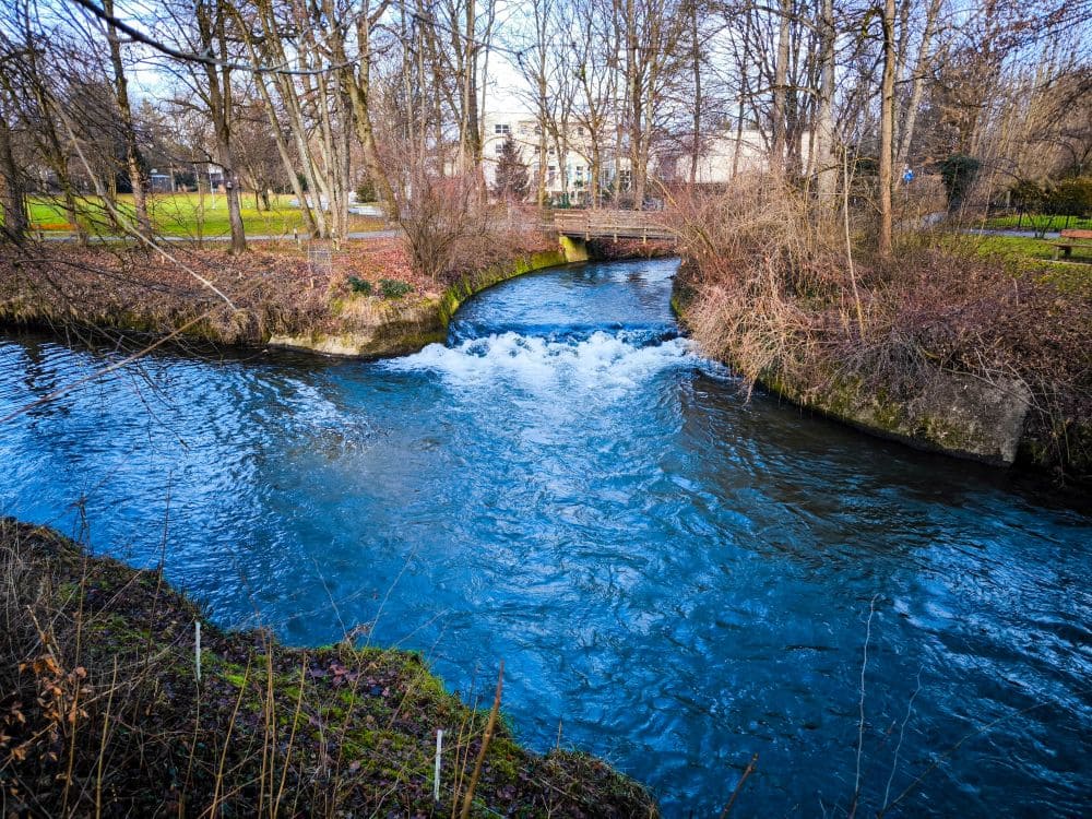 Ein leuchtend blauer Bach schlängelt sich durch die malerische Landschaft von Bad Aibling, fließt zwischen Grasufern und kahlen Bäumen hindurch und bildet kleine Stromschnellen über Felsen. In der Ferne sind unter dem klaren Himmel eine charmante Fußgängerbrücke und malerische Gebäude zu erkennen.