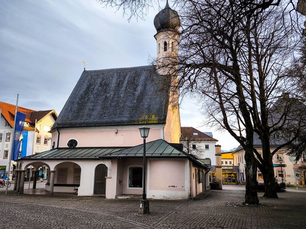 Eine kleine, historische Kirche mit einem gewölbten Kirchturm steht auf einem Kopfsteinpflasterplatz, der an den Charme des Chiemsees erinnert. Ein Baum ziert den Vordergrund, während die umliegenden Gebäude unter einem bewölkten Himmel einen Mix aus Architekturstilen zeigen.