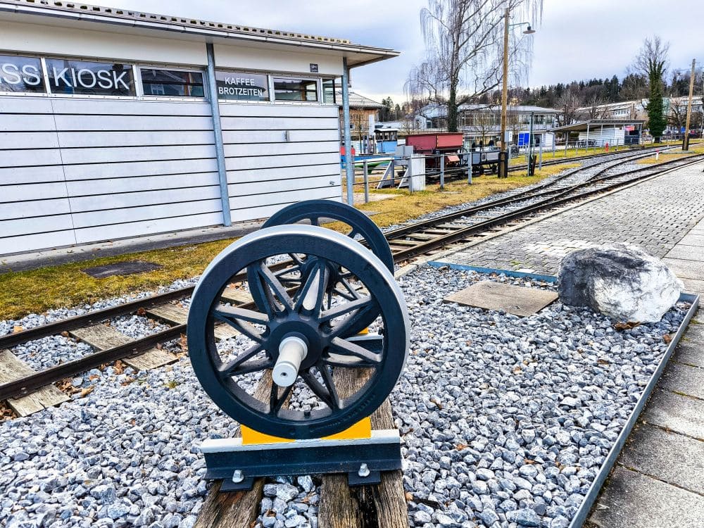 An einem malerischen Bahnhof in der Nähe des Chiemsees steht auf Schotter eine Eisenbahnradausstellung, die Gleise verlaufen parallel. Im Hintergrund sind ein Kiosk und andere Bahnhofsgebäude unter bewölktem Himmel zu sehen. Neben der Ausstellung liegt ein großer Felsen, der den malerischen Charme dieser friedlichen Seelandschaft noch verstärkt.