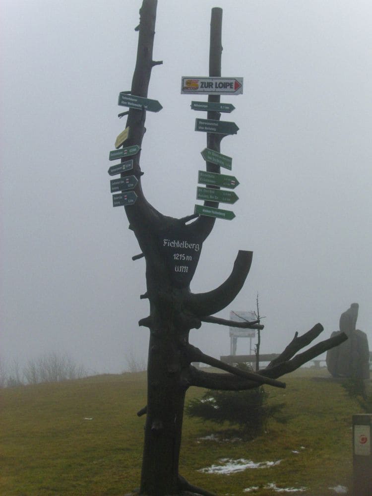 Ein zu einem Wegweiser umgearbeiteter Baumstamm steht auf dem Fichtelberg, wo bunte Wegweiser in verschiedene Richtungen weisen. Die neblige Kulisse sorgt für einen Hauch von Nebel, während der Text „Fichtelberg 1215 m üNN“ lautet und so ein immersives Erlebnis in der Graslandschaft schafft.