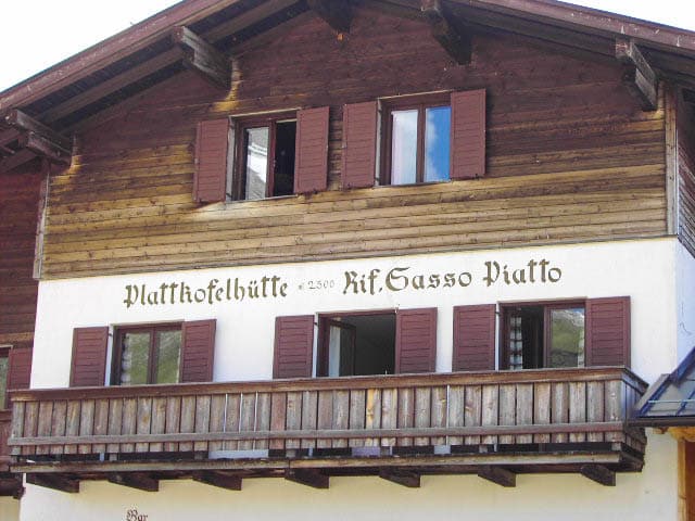 Das charmante Holzchalet Plattkofelhütte verfügt über einen Balkon mit braunen Fensterläden vor den weißen unteren Wänden. Auf der Fassade steht elegant „Plattkofelhütte 2350 Rif. Gasso Piatto“ und lädt die Gäste ein, das alpine Ambiente zu genießen.
