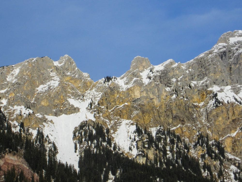 Die zerklüftete Bergkette im Tannheimer Tal besticht durch schneebedeckte Gipfel und Klippen unter einem strahlend blauen Himmel. Hohe Nadelbäume säumen die unteren Hänge und bilden einen Kontrast zum felsigen Gelände darüber. Perfekt für eine winterliche Wanderung mit Blick auf den glitzernden Haldensee.