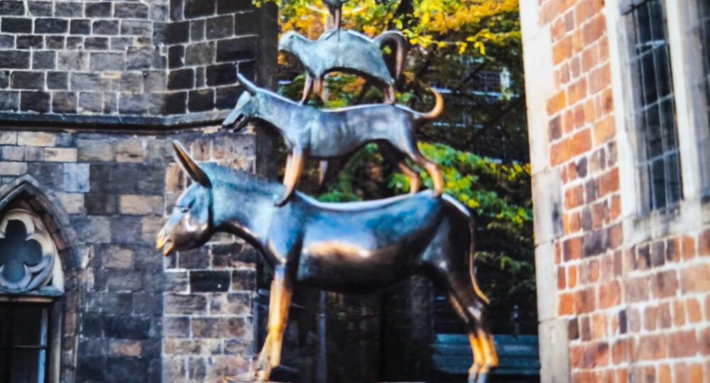 Entdecken Sie die Bremer Bronzestatue mit vier übereinander gestapelten Tieren – einem Hahn auf einer Katze, einem Hund auf einem Esel und einem Hund auf einem Esel. Eingebettet zwischen Backstein- und Steingebäuden und üppigem Grün ist diese einzigartige Sehenswürdigkeit ideal für Entdeckungstouren und Reisetipps.
