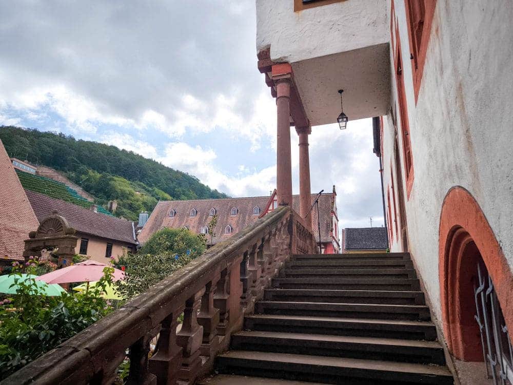 Eine Außentreppe mit verziertem Geländer führt zu einem Gebäude in Klingenberg, das durch seine markante rot-weiße Architektur besticht. Im Hintergrund bildet eine hügelige Landschaft mit Gebäuden einen Kontrast zum bewölkten Himmel und schafft eine malerische Szenerie.