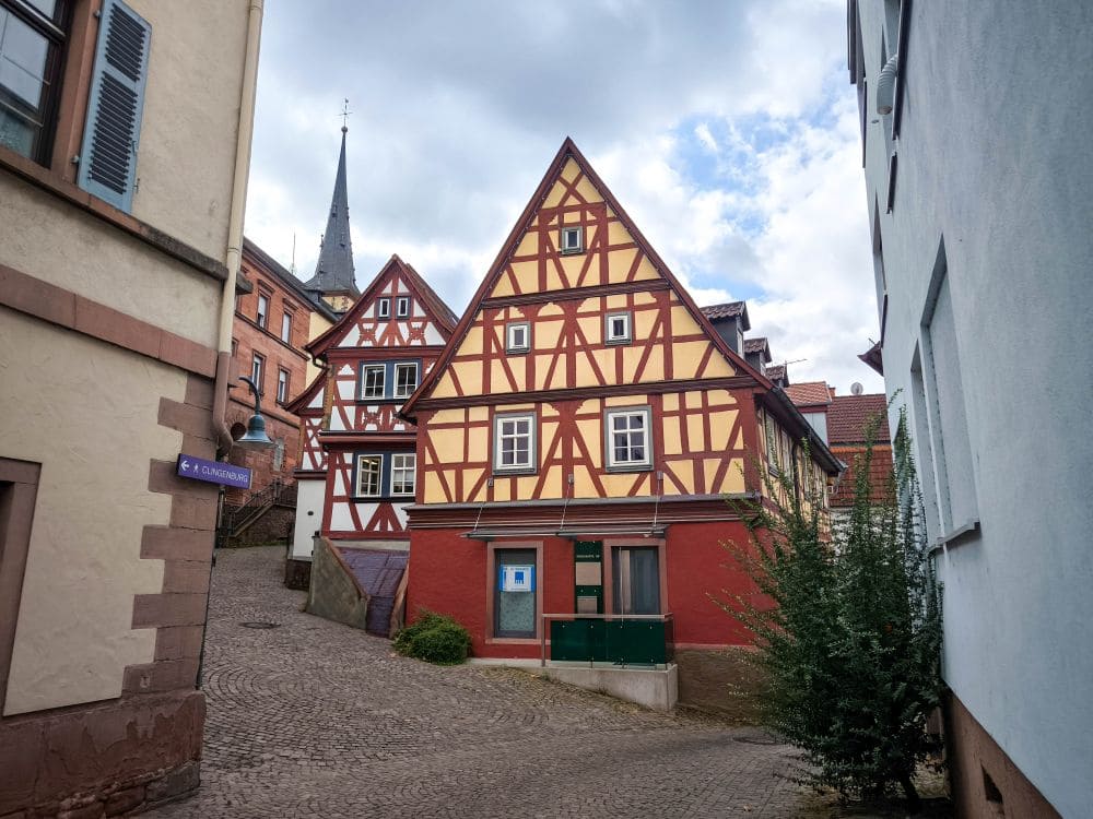 Eine Kopfsteinpflasterstraße im malerischen Dorf Klingenberg, gesäumt von traditionellen Fachwerkhäusern mit roten und gelben Fassaden. Ein Kirchturm ragt in den leicht bewölkten Himmel, während links ein Straßenschild den Weg durch dieses charmante europäische Juwel weist.