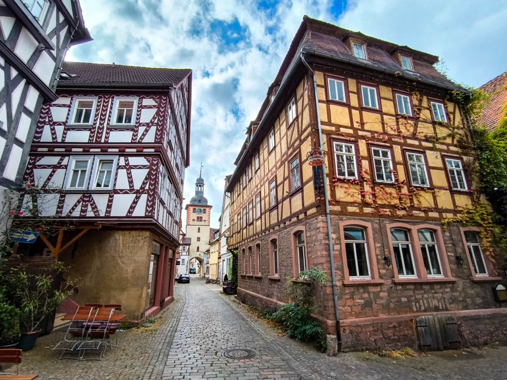 Eine malerische Kopfsteinpflasterstraße im idyllischen Europastädtchen Klingenberg, gesäumt von Fachwerkhäusern. Der Himmel ist bedeckt, im Hintergrund ist ein Kirchturm zu sehen. Links stehen ein paar Topfpflanzen und Sitzgelegenheiten im Freien.
