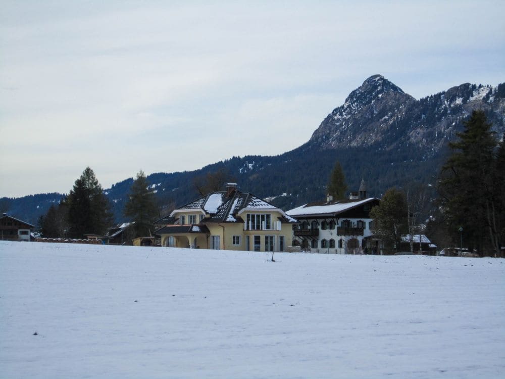 Inmitten einer schneebedeckten Landschaft liegt das große zweistöckige Haus mit gelb-weißen Wänden und dunkel getönten Fenstern eingebettet zwischen Bäumen. Im Hintergrund erheben sich majestätisch die Berggipfel des Tannheimer Tals unter einem wolkigen Himmel und vermitteln den ruhigen Charme einer winterlichen Wanderung in der Nähe des Haldensees.