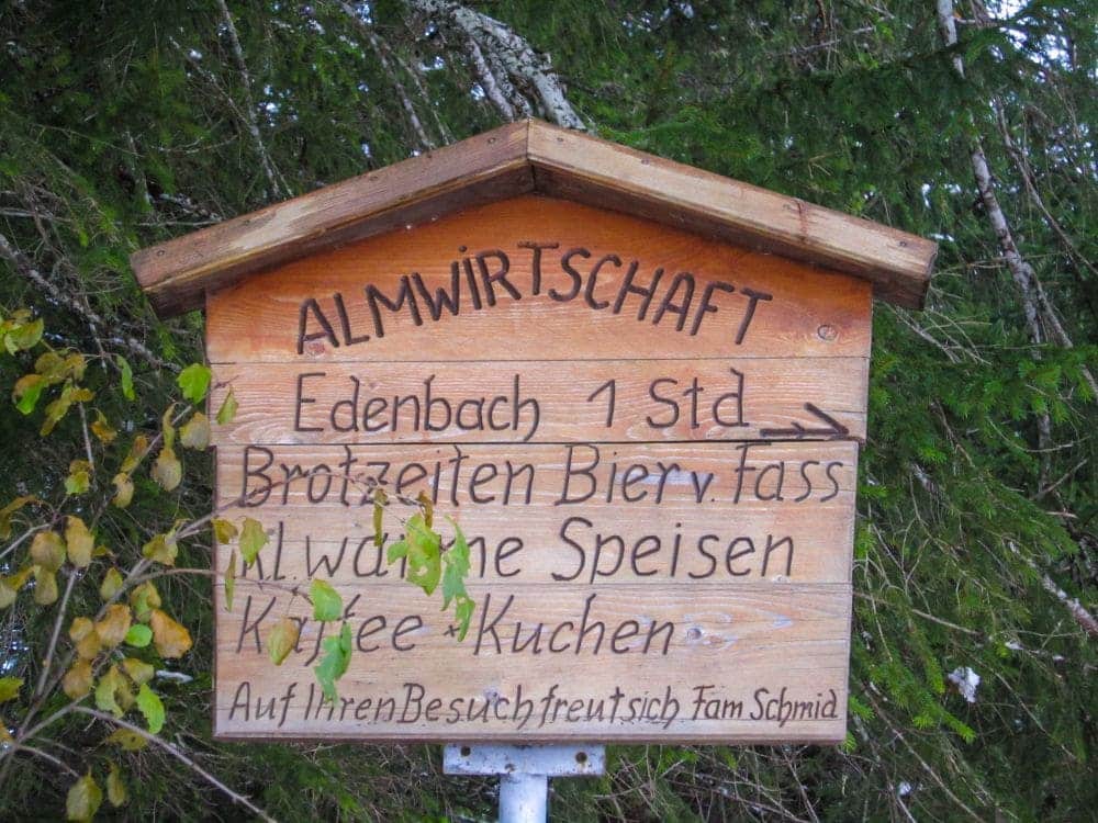Das Holzschild mit der Aufschrift „Almwirtschaft Edenbach 1 Std.“ lädt Wanderer während ihrer winterlichen Wanderung im Tannheimer Tal zu einer gemütlichen Rast ein. Genießen Sie Snacks, Bier vom Fass, warme Gerichte, Kaffee und Kuchen. Familie Schmid heißt Sie herzlich willkommen inmitten der grünen Natur am Haldensee.