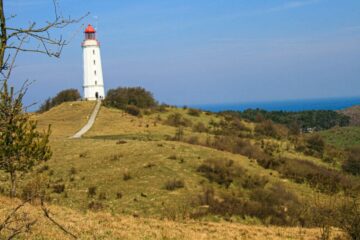 Ein hoher weißer Leuchtturm mit roter Spitze steht auf einem grasbewachsenen Hügel auf Hiddensee. Ein schmaler Pfad führt hinauf, umgeben von spärlichen Bäumen und Sträuchern. Im Hintergrund erstreckt sich das blaue Meer unter einem klaren Himmel und bietet einen ruhigen Küstenblick.