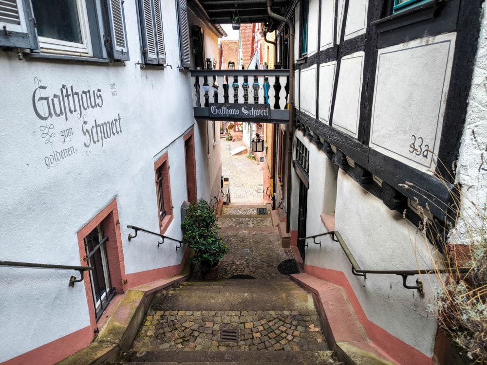 Eine schmale, kopfsteingepflasterte Gasse mit Stufen schlängelt sich durch Klingenberg, gesäumt von traditionellen Fachwerkhäusern. An der linken Wand ist „Gasthaus Schwert“ aufgemalt. Ein Balkon mit Holzgeländer bietet einen schönen Ausblick auf die malerische Dorfstraße.
