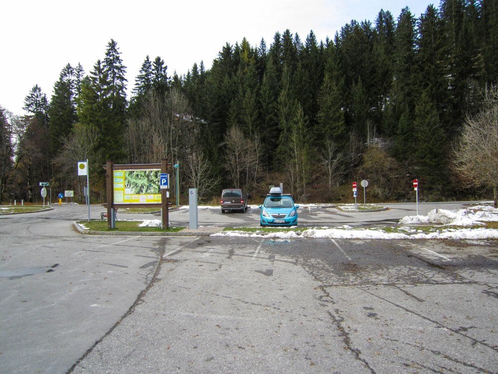 Ein fast leerer Parkplatz mit zwei Autos in der Nähe einer großen Informationstafel lädt zu einer winterlichen Wanderung ein. Dahinter ist der dichte Wald aus immergrünen Bäumen, der an das Tannheimer Tal erinnert, mit Schnee bedeckt. Der Himmel ist weiterhin bedeckt, was die ruhige Landschaft noch verstärkt.
