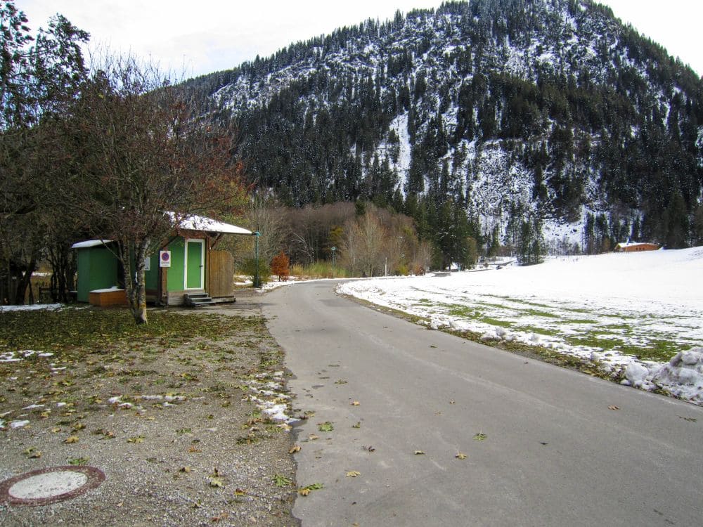 Eine Bergstraße schlängelt sich durch die verschneite Landschaft im Tannheimer Tal. Links steht ein kleines grünes Gebäude mit teilweise schneebedecktem Dach. Bäume säumen die Straße, und im Hintergrund ragt unter einem wolkigen Himmel ein schneebedeckter Berg empor und lädt Winterwanderer zu einer Wanderung ein.