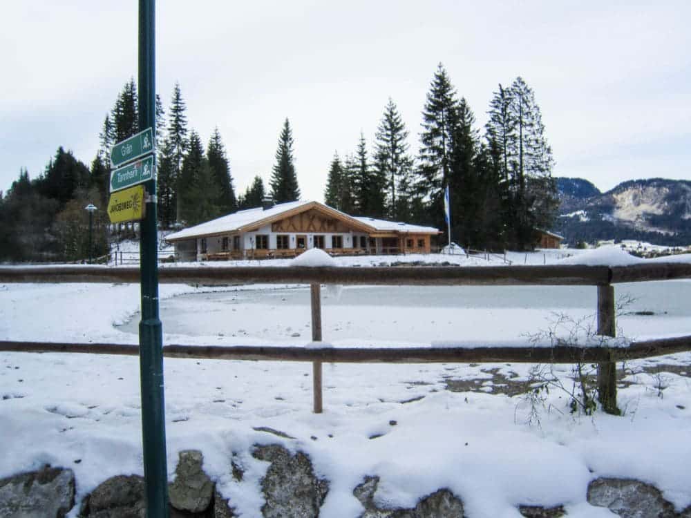 Eine verschneite Landschaft zeigt eine von Kiefern umgebene Holzhütte im bezaubernden Tannheimer Tal. Im Vordergrund liegt der zugefrorene Haldensee, eingefasst von einem rustikalen Zaun. Links steht ein Straßenschild mit mehreren Richtungsangaben unter dem bewölkten Himmel, mit majestätischen Bergen im Hintergrund – perfekt für eine winterliche Wanderung.