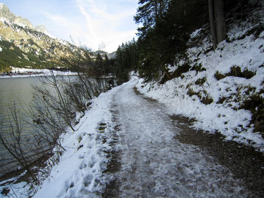 Eine winterliche Wanderung entlang des schneebedeckten Pfades am Haldensee, eingebettet in einen ruhigen Wald auf der einen Seite und der majestätischen Kulisse der Berge des Tannheimer Tals unter einem strahlend blauen Himmel.