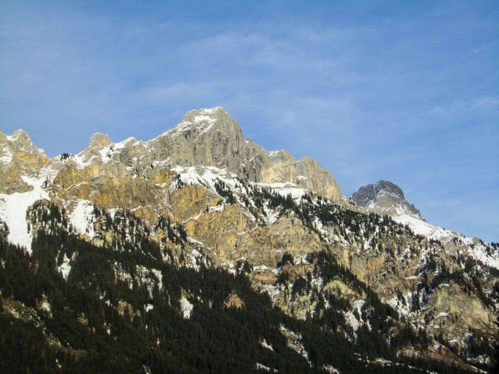 Schneebedeckte Bergketten mit felsigen Gipfeln unter einem klaren blauen Himmel erinnern an das malerische Tannheimer Tal. Die unteren Hänge sind mit dichten immergrünen Wäldern bedeckt, perfekt für eine winterliche Wanderung.