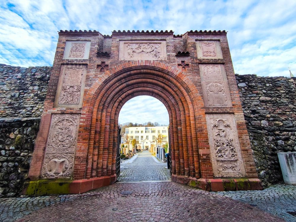 Ein historischer Torbogen aus Ziegelstein und Stein mit kunstvollen Schnitzereien sticht unter einem blauen, teilweise bewölkten Himmel hervor. Durch den Bogen führt ein gepflasterter Weg zu einem hellen Gebäude in Wasserburg am Inn, flankiert von kleineren Gebäuden voller Geschichte und Genuss.