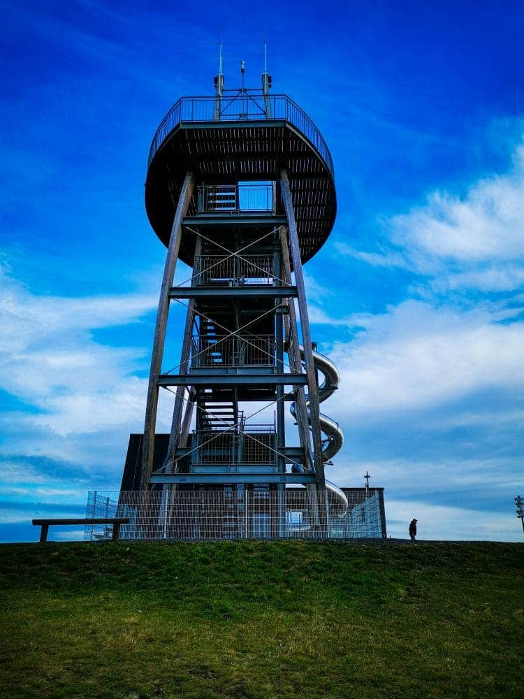 Noahs Segel, ein hoher Aussichtsturm mit Spiralrutsche, steht stolz auf einem grasbewachsenen Hügel unter einem strahlend blauen Himmel mit vereinzelten Wolken. Eine Person in der Nähe betont die beeindruckende Höhe des Bauwerks.